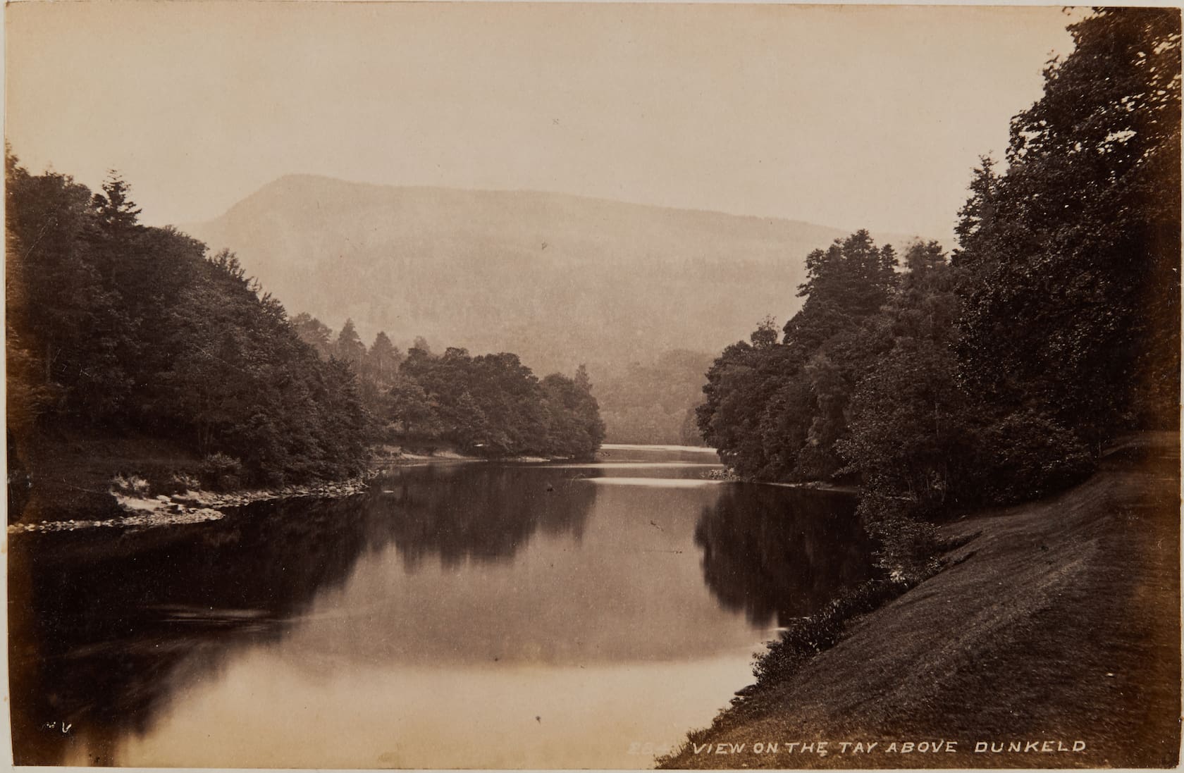A sepia photograph of a calm river with forested banks and misty hillsides in the background.