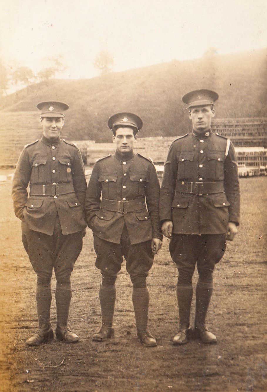 A sepia photograph of 3 men in uniform standing casually on grass, with stacked wood behind them.
