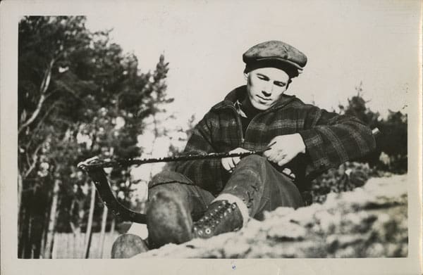 A black and white photograph of a young man in plaid jacket and peaked cap. He sits on the ground, sharpening a blade braced against his knee.