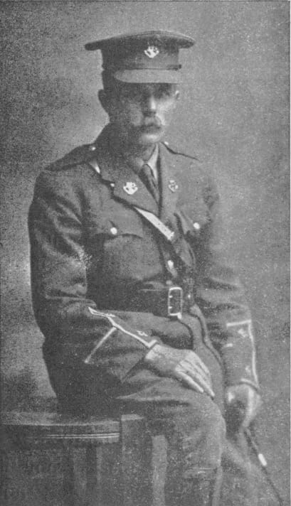 A black and white photograph of a middle-aged man with handlebar moustache, in full uniform and cap. He poses with one hip resting on a table, hand on his right knee.