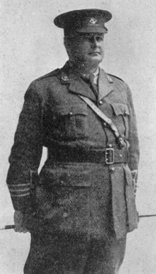 A black and white photograph of a middle-aged man in full belted uniform and cap. He stands erect, both hands holding his swagger stick horizontally behind him.