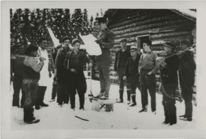 A black and white photograph of a uniformed man standing atop a wooden crate outside two log buildings. He reads from a newspaper to a dozen lumber workers standing in snow around him.