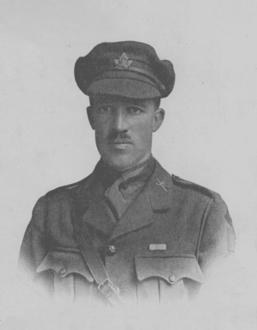 A black and white, head-and-shoulders photograph of a moustached man in uniform wearing a cap emblazoned with a maple leaf badge. He stares unsmiling at the camera, head tilted slightly down.