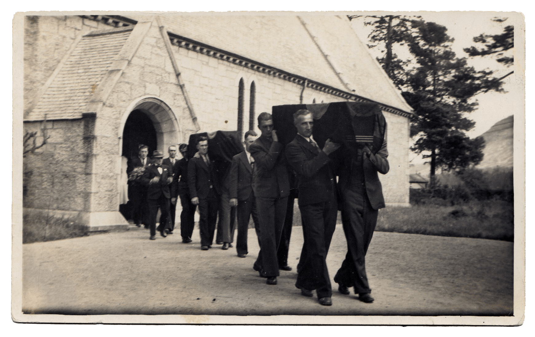 A sepia photograph of a dozen unsmiling men in suits leading a procession out of the side door of a stone church. They support two flag-draped coffins on their shoulders.