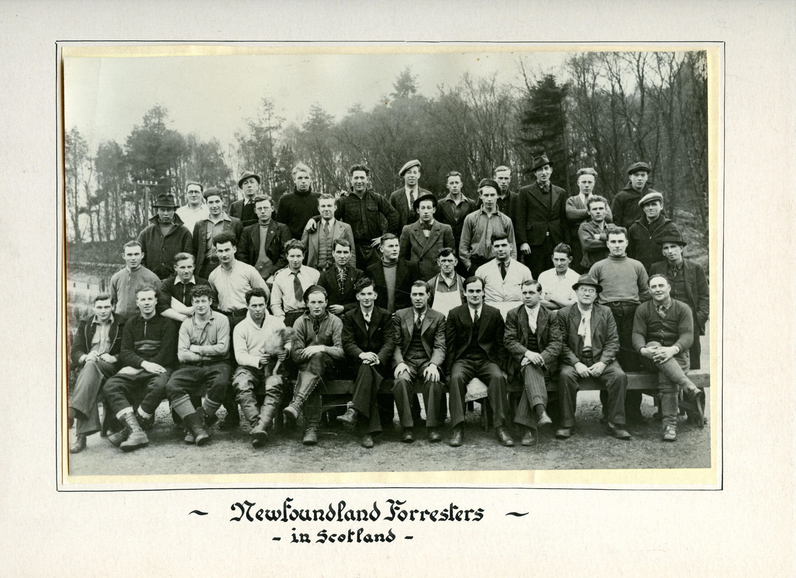A black and white photograph of 40 men dressed casually or in suits. They sit or stand in 4 rows, posed for an outdoor photo. Many men smile and look at the camera. A row of trees is in the distance behind them.