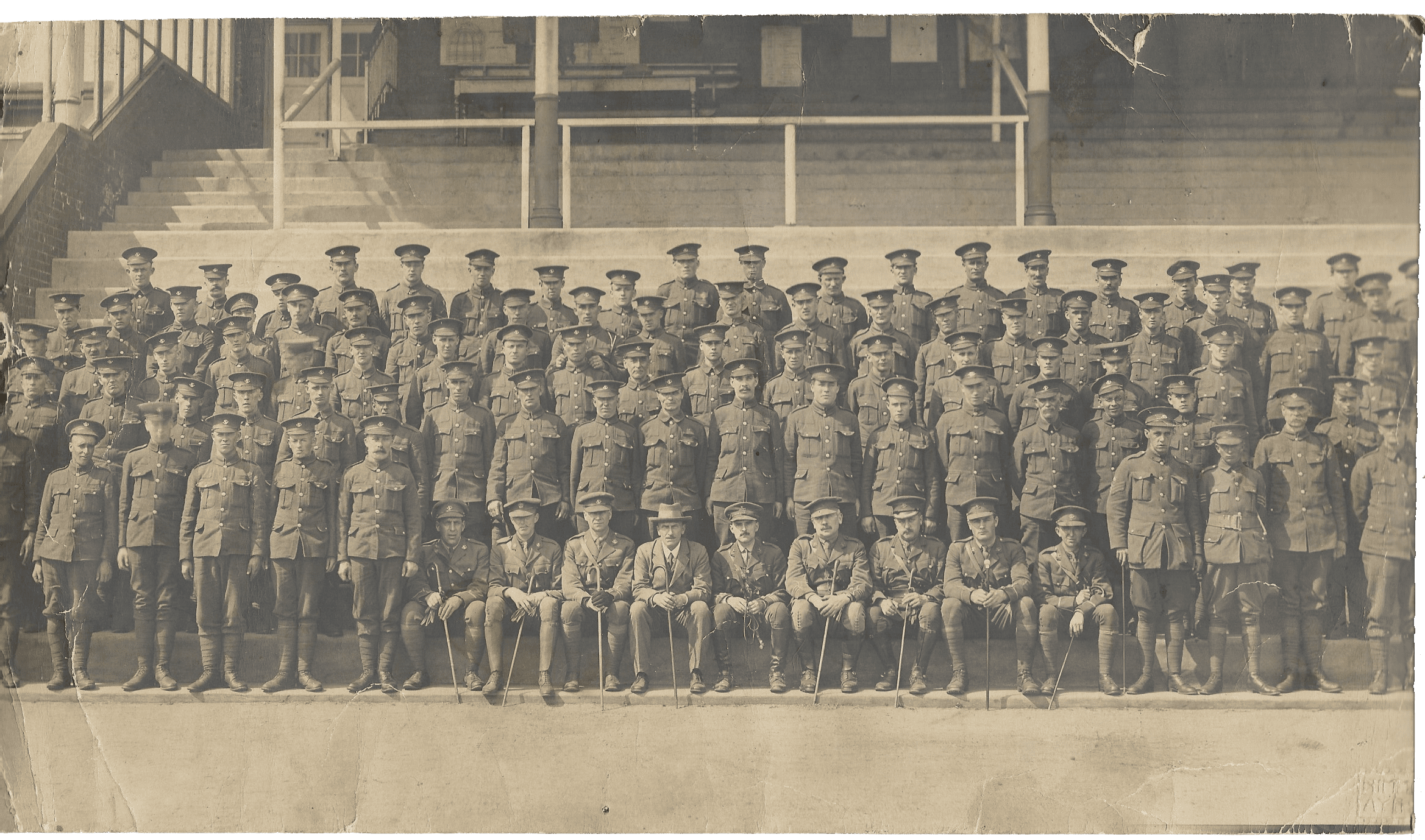 A sepia photograph of 5 rows of uniformed soldiers standing at attention on or before concrete benches. Seated in the centre of the first row are 9 officers and one civilian.