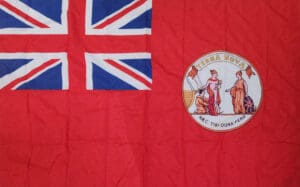 A colour photograph of a bright red flag in excellent condition. In its top left corner, taking up a quarter of the flag area, is a Union Jack. To its right, centered vertically, is a circle badge that features 3 figures (a fisher, the god Mercury, and Britannia) and above them the words Terra Nova in gold print.