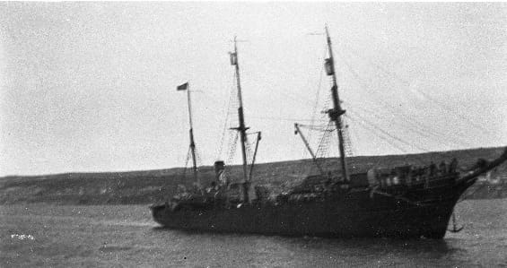 A somewhat grainy black and white photograph of a three-masted vessel with a high bow and bowsprit, taken as it steams up a wide harbour. The land on the far side is visible but its features are not.