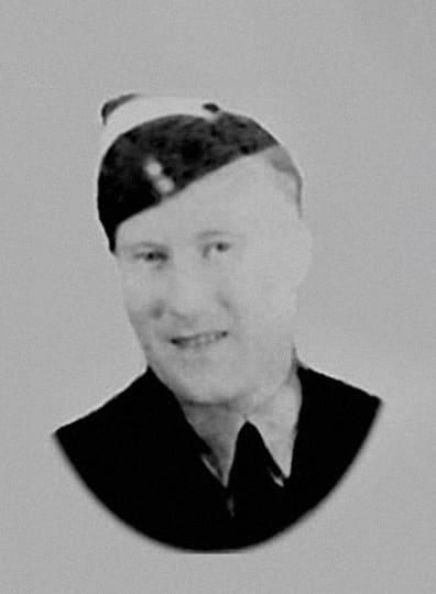 A black and white, head-and-shoulders photograph of a fair-haired smiling man in uniform. His head and wedge cap are tilted to the left.