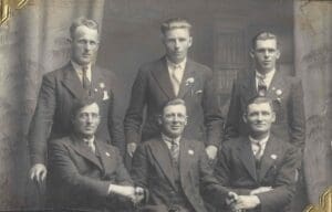 A black and white studio photograph of 6 men in suits and ties with N O F U pins on their lapels. Three men stand in back and the other three are seated in front of them. The man sitting in the middle holds one hand of each man beside him.