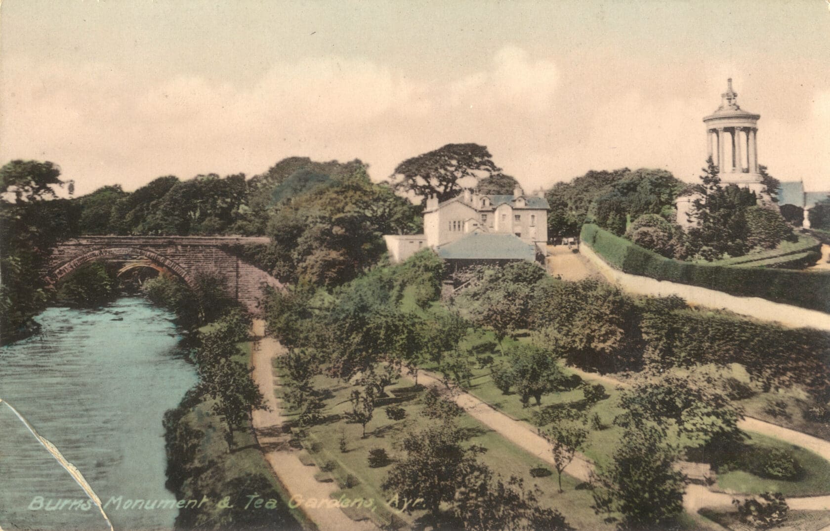 A colorized photograph of a large white building and well-tended gardens above the right-hand bank of a river. A dark stone bridge spans the waterway and a tall circular monument with several columns rises across a road from the building. 
