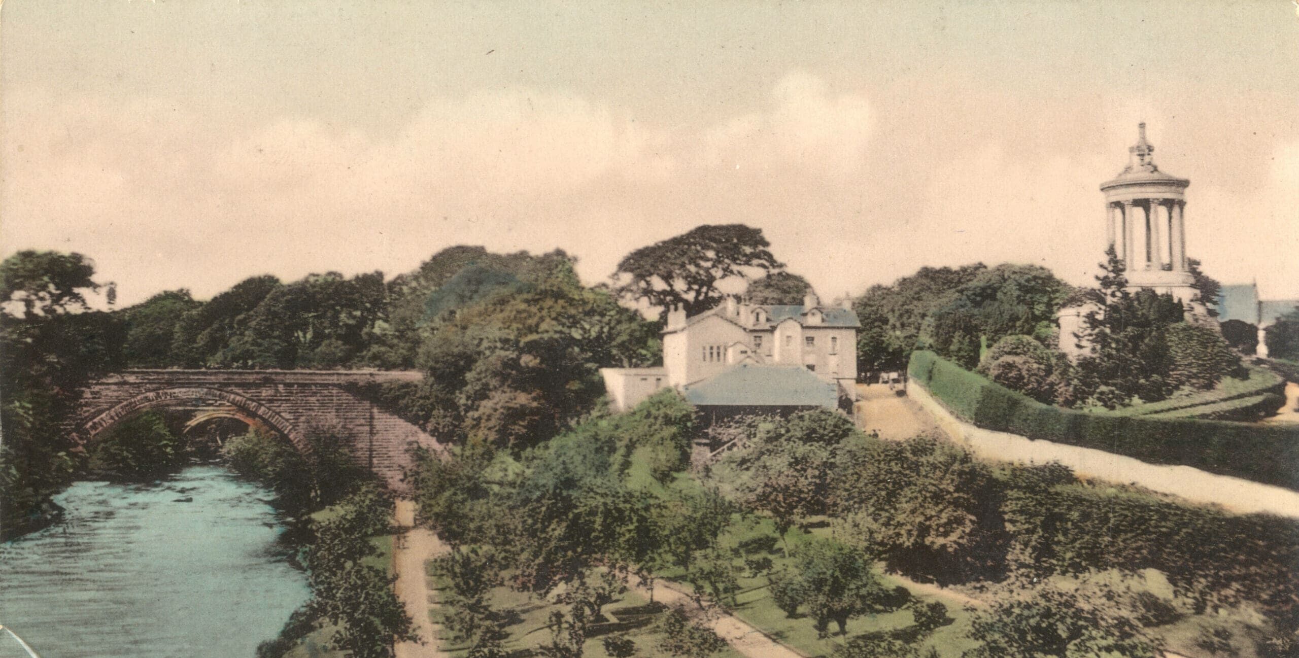 A colorized photograph of a large white building and well-tended gardens above the right-hand bank of a river. A dark stone bridge spans the waterway and a tall circular monument with several columns rises across a road from the building.