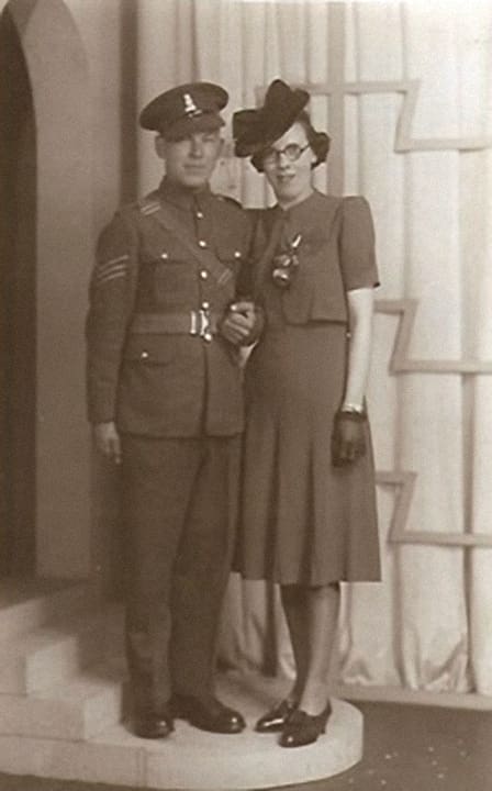 A sepia photograph of a uniformed soldier and a woman in hat, glasses, short-sleeved dress and gloves. They stand on a step below a doorway and in front of a curtain, smiling and holding hands.