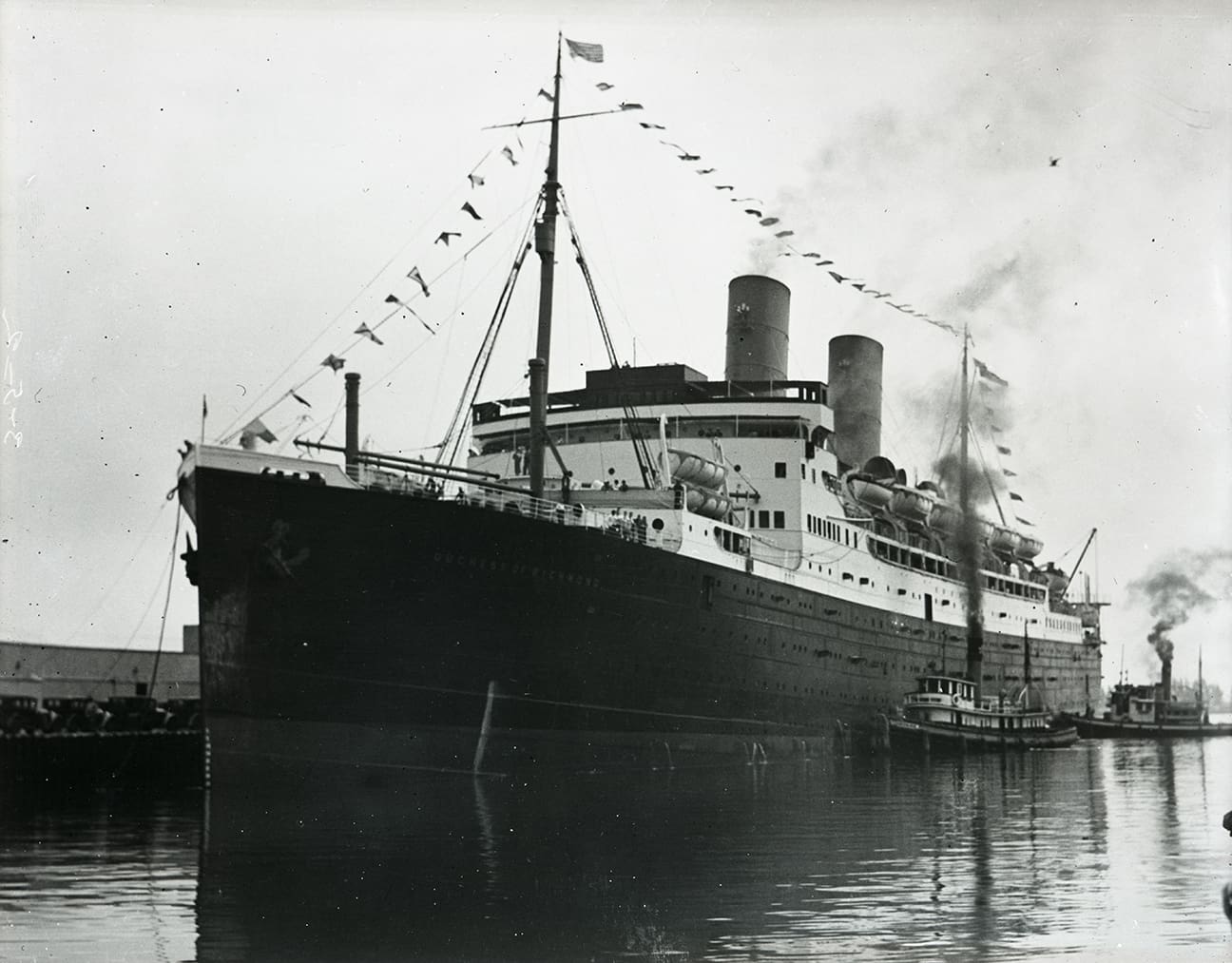 A black and white photograph of the bow and port side of a dark-hulled liner with two tugboats alongside. The ship is dressed with flags.