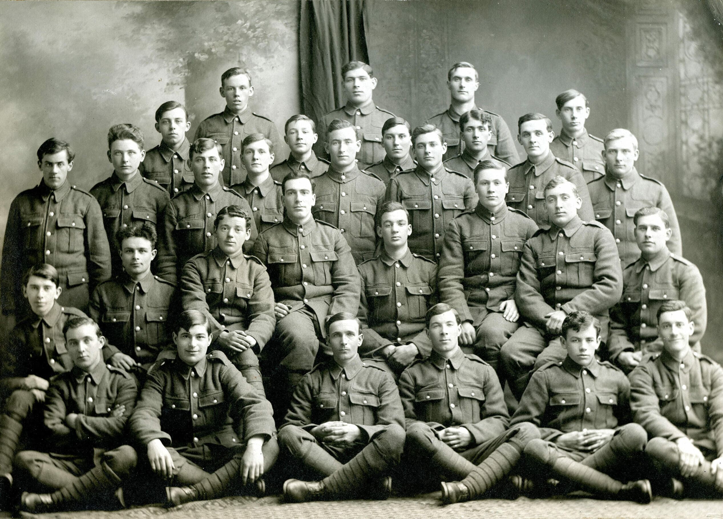 A black and white studio photograph of 30 uniformed men in 5 rows. Those in the first row sit cross-legged on carpet. The men have varied expressions and look straight at the camera.