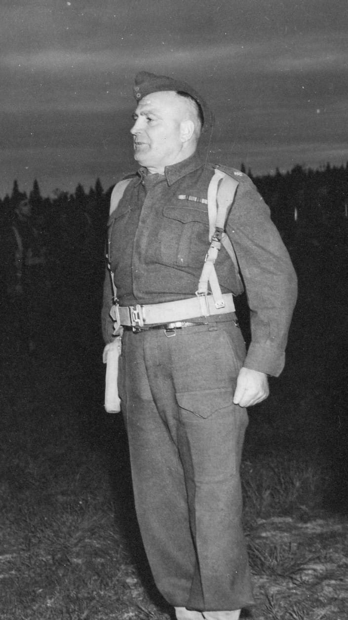 A black and white photograph of a middle-aged man in uniform. He stands at attention on grass facing left with his left fist clenched. A night sky and dark forest are visible beyond him.
