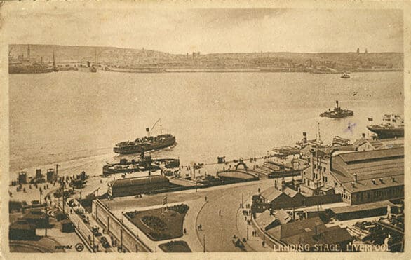 A sepia aerial photograph of a wide river and activity on its nearest shore. On the broad riverside apron people mill about. Tidy buildings, streets and parks flank the waterfront. Small ships are docked or steaming past.