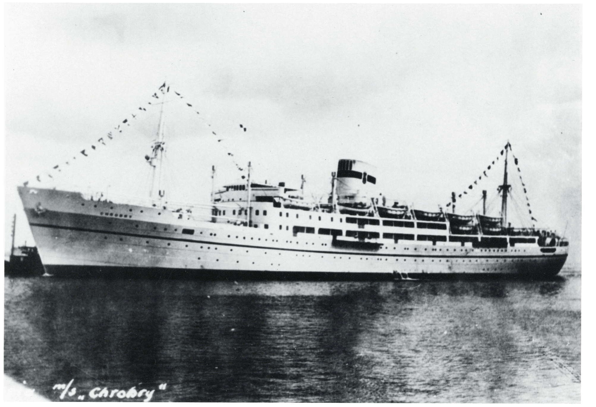 A black and white photograph taken looking down on the open deck of a ship that is thronged with men wearing caps and life jackets. Many of them look up at the camera.