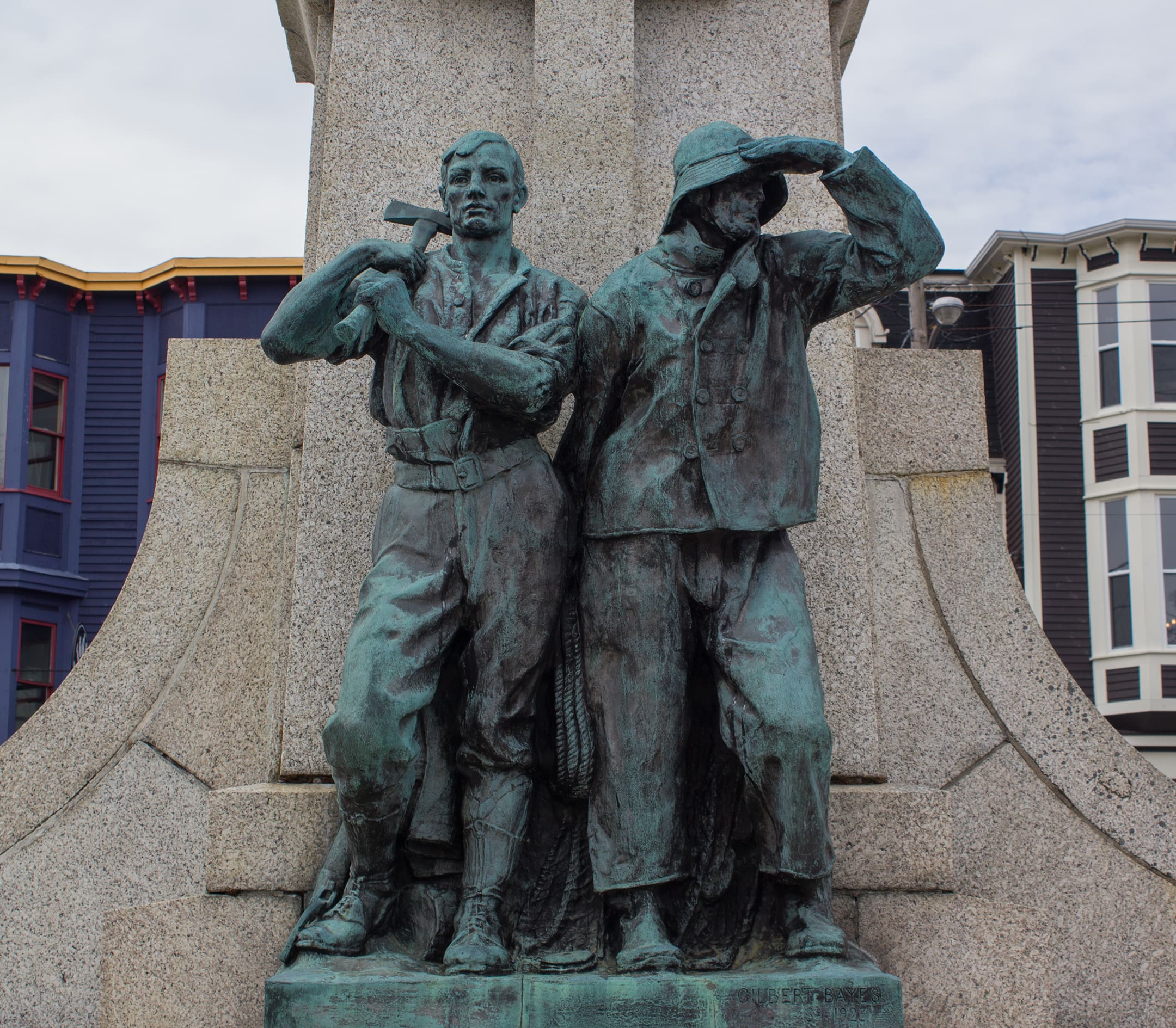 A modern-day colour photograph of two weathered bronze statues. The figure of the forester is at left. His sleeves are rolled up and an axe rests on his shoulder. At right, a mariner in oilskins holds his hand to his sou’wester brim and scans the horizon.