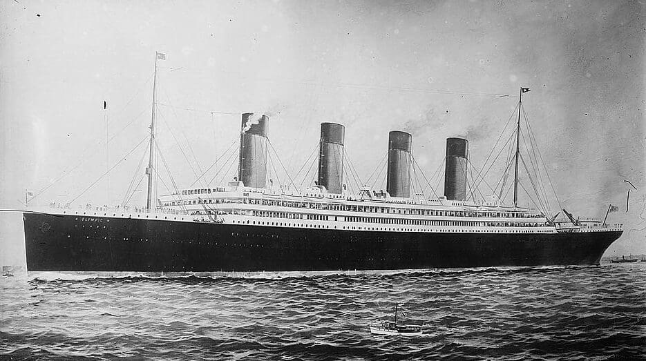 A black and white drawing of a long ship with 4 funnels. It has a dark hull and a three-storey white superstructure. It is depicted steaming to the left past a small fishing vessel in the foreground. 