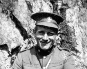 A black and white head and shoulders photograph of a man with a warm, wide smile and square jaw. He wears a uniform and cap. Behind him is a rough cliff face. 