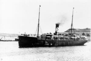A black and white photograph of a dark-hulled steamship seen from the port side. It has a mast fore and aft, a funnel amidships, and a row of lifeboats on davits outside the superstructure. It is steaming away from a grassy shore dotted with a few buildings.