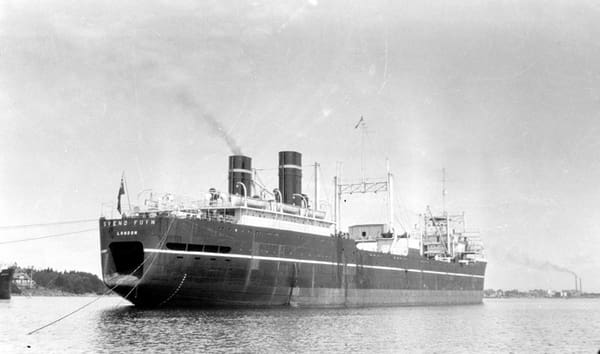A faded black and white photograph of the aft end and starboard side of a dark-hulled ship at anchor. It rides high on its waterlines and stern cables run aft into the water. A low shoreline lies behind.