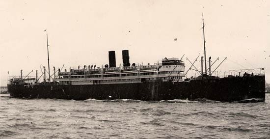 A dark-hulled ship with two funnels amidships and loading cranes fore and aft. It steams to the right in choppy waters.