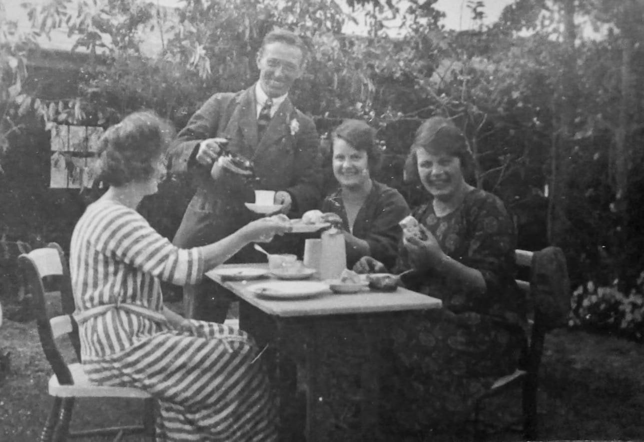 A black and white photograph of a man in a suit pouring tea for three women seated at an outdoor table. All are smiling, three of them directly at the camera.