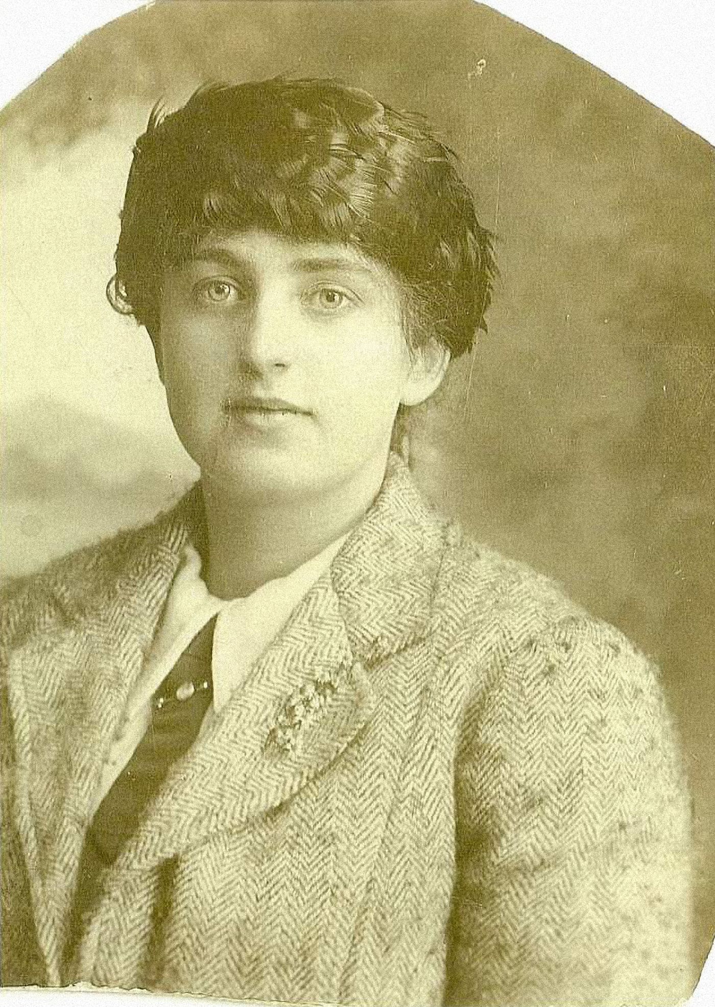 A sepia studio photograph of a woman in a close-fitting hat of feathers over hair pulled back, a thick herringbone tweed jacket, a white shirt and tie. Her oval face is distinguished by large pale eyes, a clear complexion and full lips.