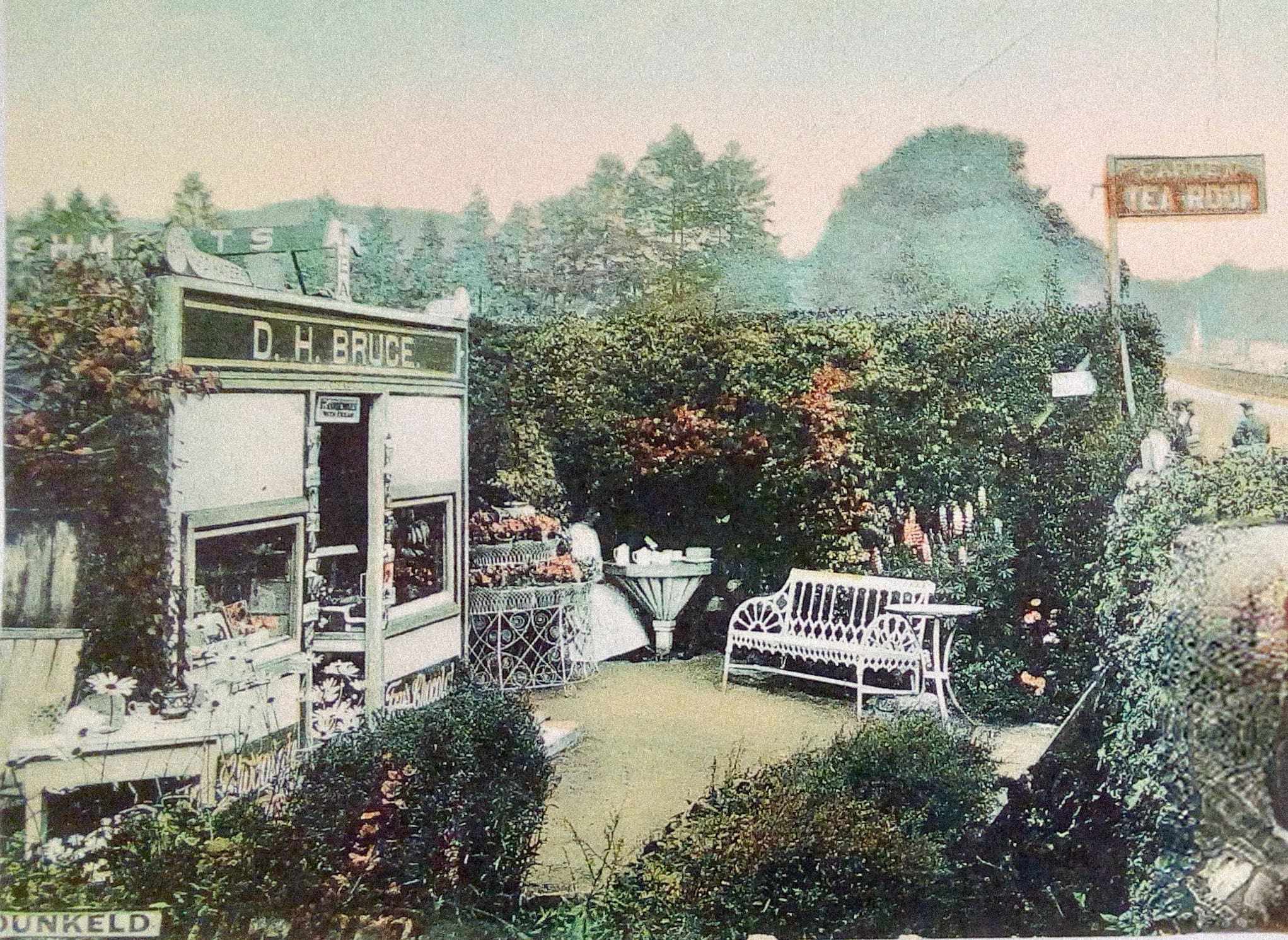 A colourized photograph of a small green area enclosed by high hedges. On the left is a storefront. A large sign over the door reads D.H. Bruce. The doorway is flanked by large windows. Around the green area are a bench, two tables with a tea service laid out, and many plantings.