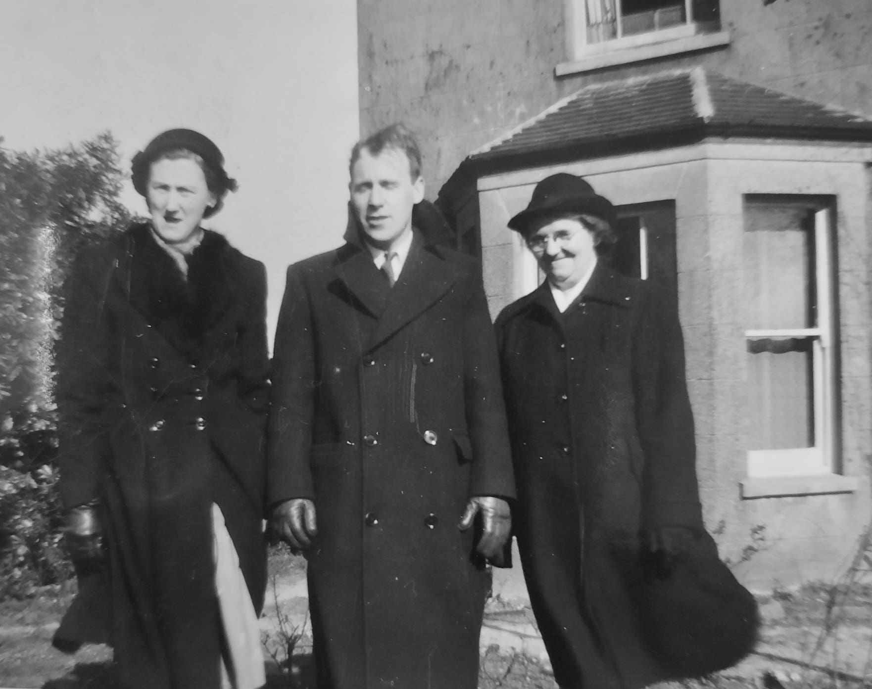 A black and white photo of three people standing abreast in the front yard of a house. The wind blows their long dark overcoats. The women wear hats and the man has leather gloves on.