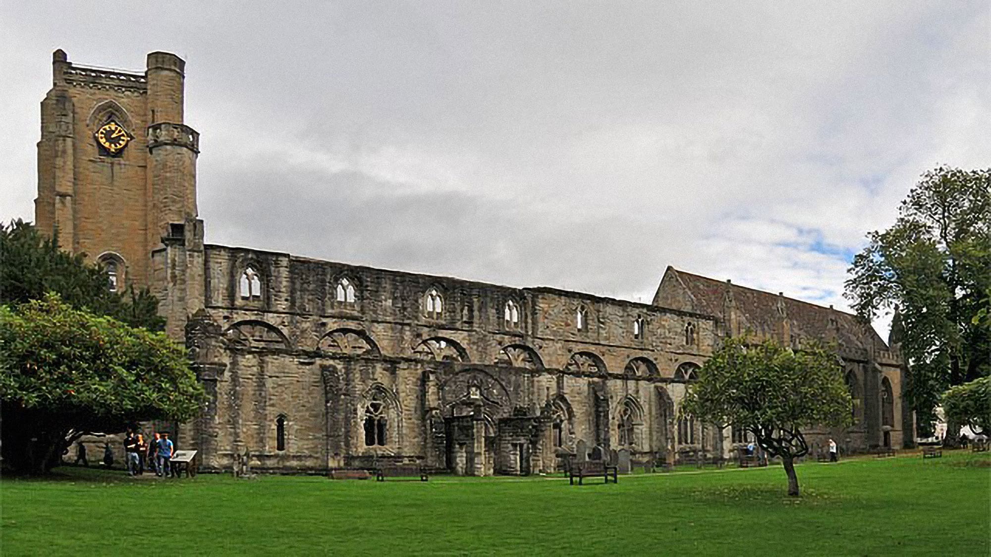 A well-tended green lawn in the foreground leads to long, grey side walls of the cathedral’s ruined stone nave. A tall stone tower on the left has a clock with gold numerals. To the right of the nave, the roof and shape of the actual cathedral are visible.