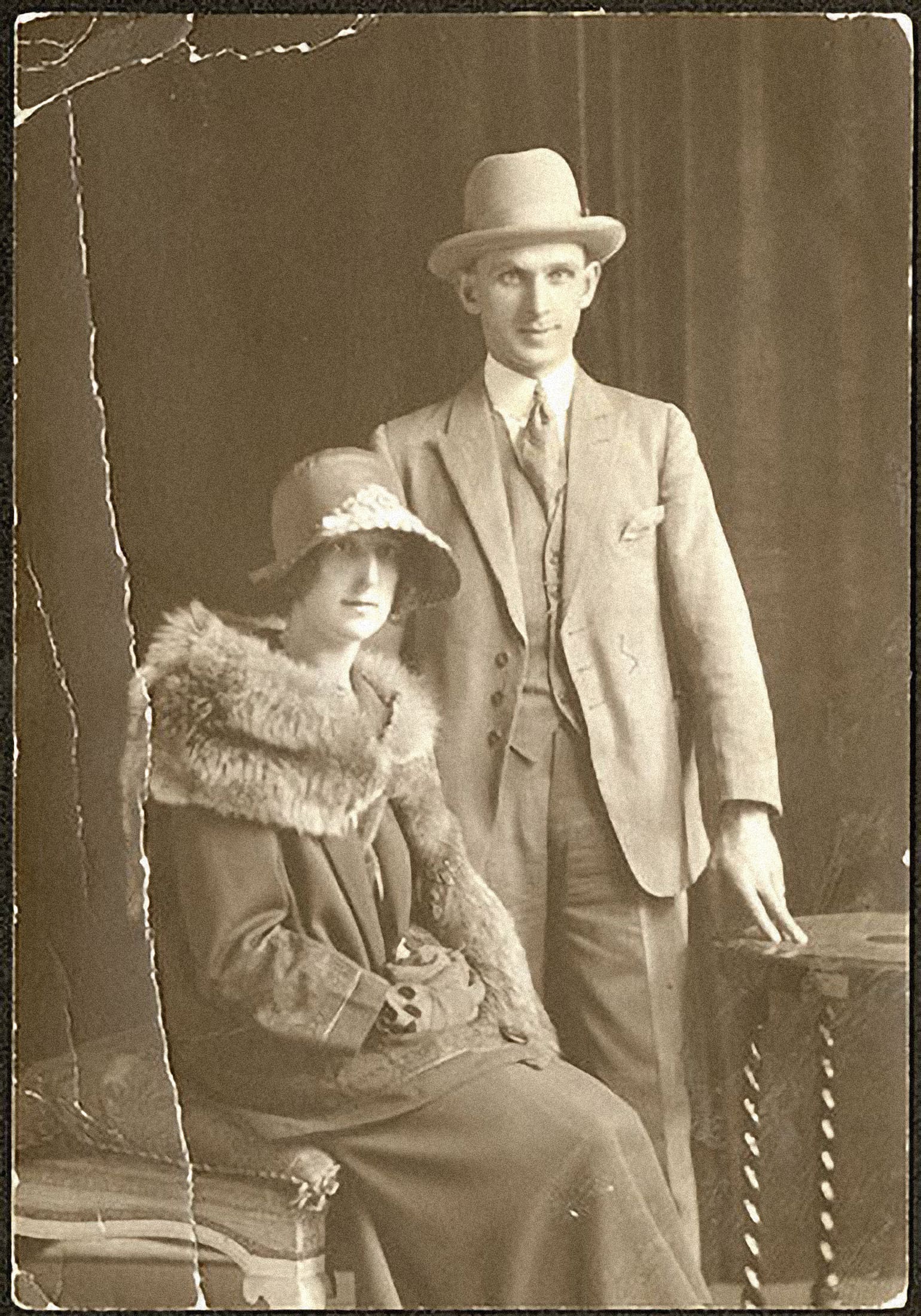 A sepia studio photograph of a man and a woman. She sits at left, wearing woollen clothes, gloves, a large hat, with a fur around her shoulders. He stands to her right, two fingers outstretched to touch a small table. He wears a light-coloured, three-piece suit, high-collared shirt with a tie, and a fedora.