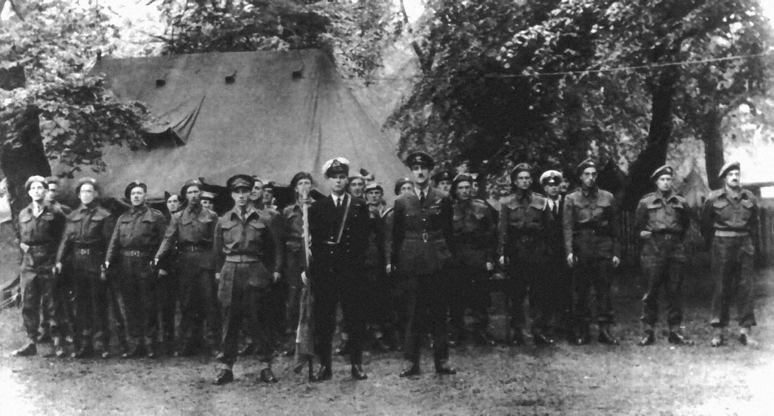A black and white photo of men in uniform who are standing at attention, rifles by their sides, in front of a tent and some leafy trees. 