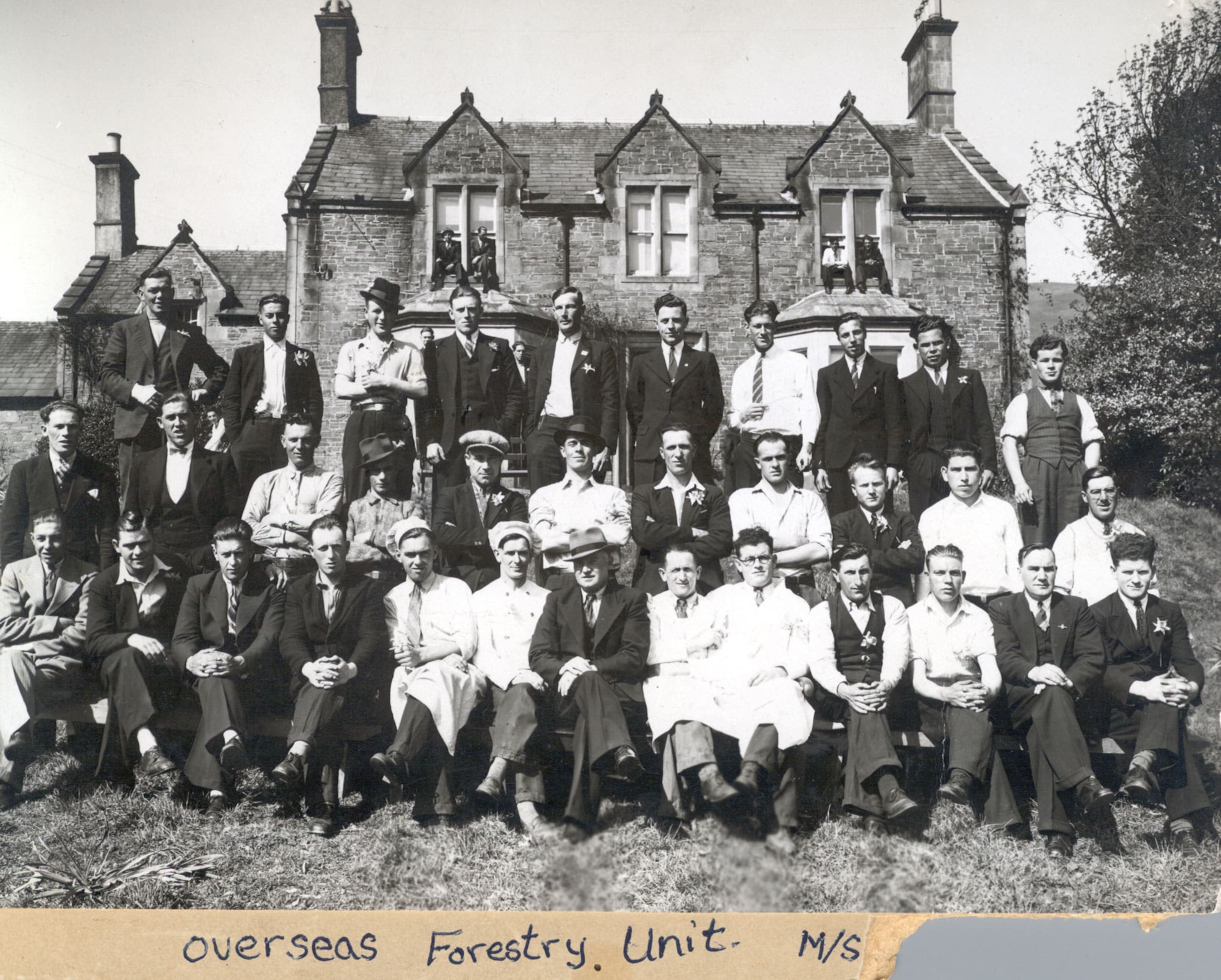 A black and white photograph of three dozen men, many in suits and ties, posing in three rows in front of a stone house. A few more men sit in second-storey windowsills.