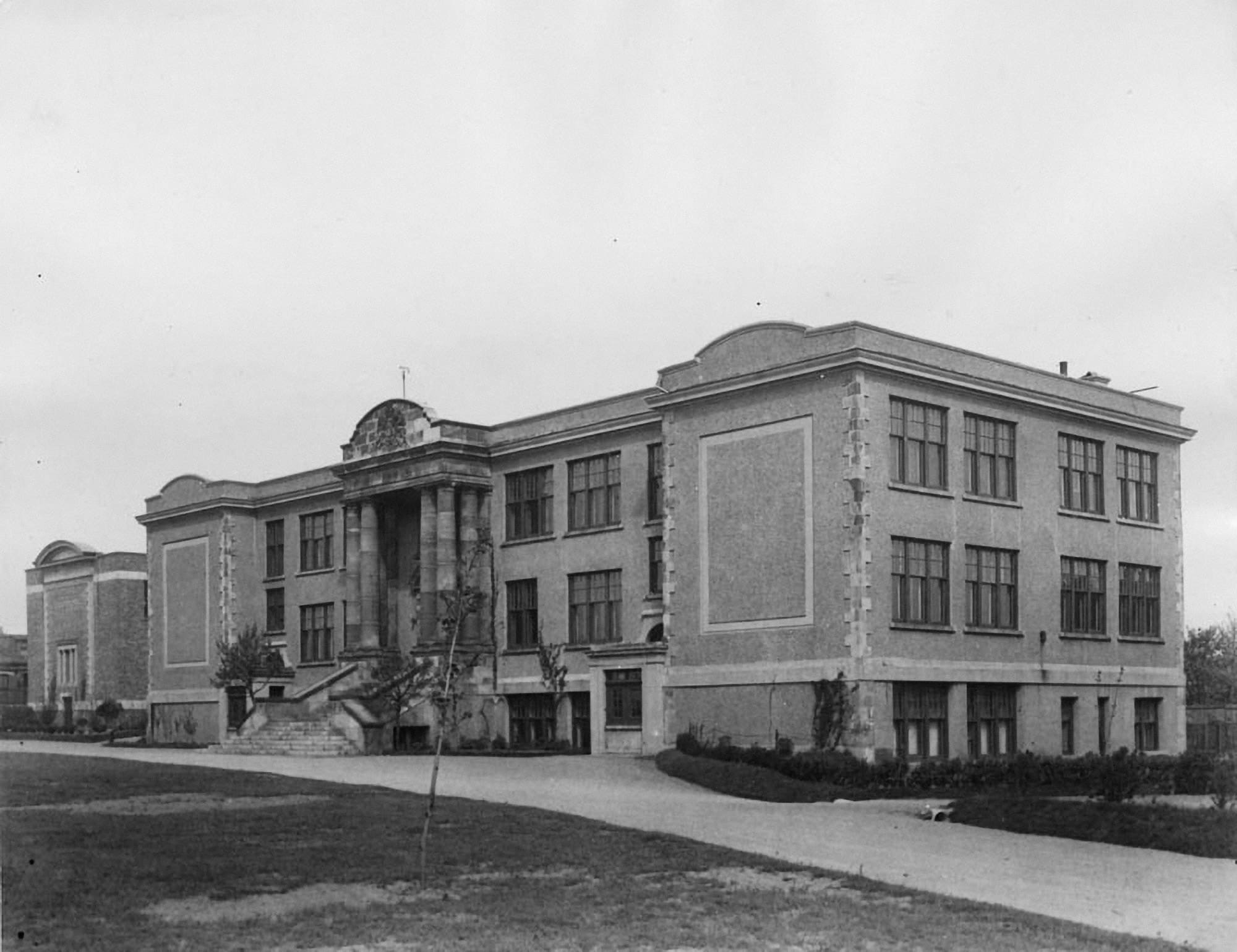 A black and white photograph of a solid-looking, two-storey brick building with rows of tall windows. The front door has a stone portico extending to the roofline. Several stone columns rise each side of the doorway.