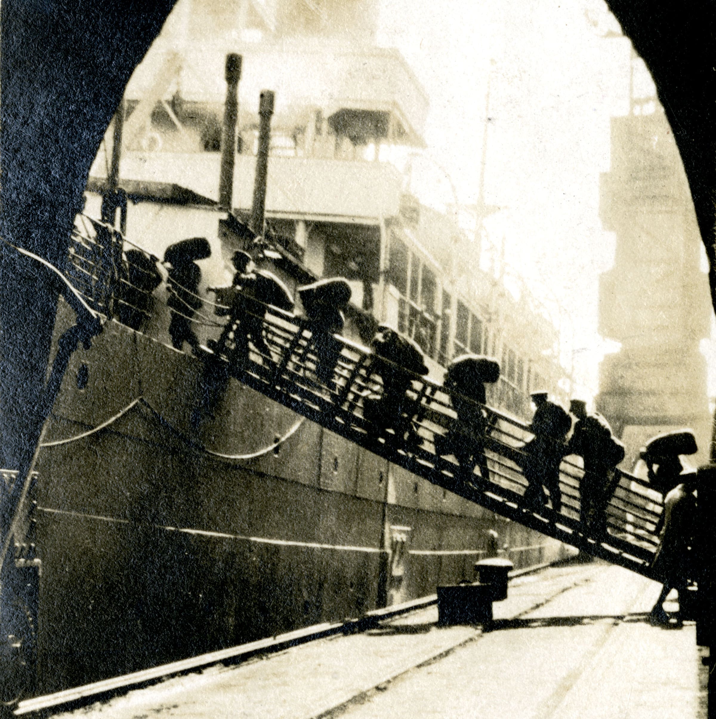 A sepia photograph of soldiers carrying sacks over their shoulders walking single file up a gangplank and onto a ship. They are silhouetted against a bright background. Railway tracks on the brick embankment run under the gangplank and into the distance.