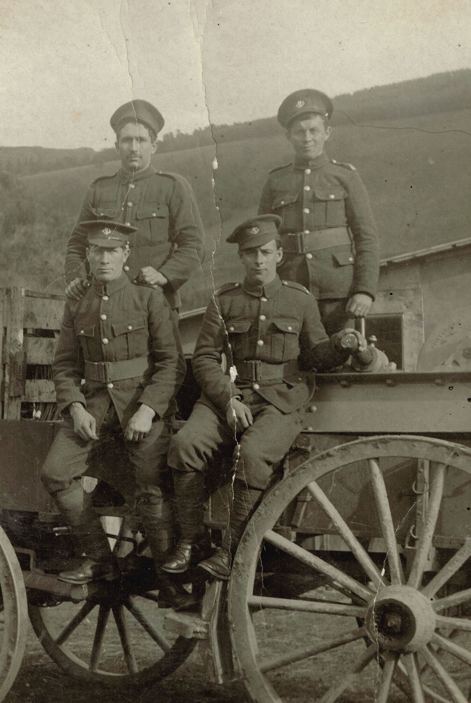 A sepia photograph of four men in uniforms and caps. Two are perched on the edge of a wagon bed, one with his hand resting on a piece of equipment. The other two stand in the wagon behind them. A bare hill with forest at its upper levels rises behind them.