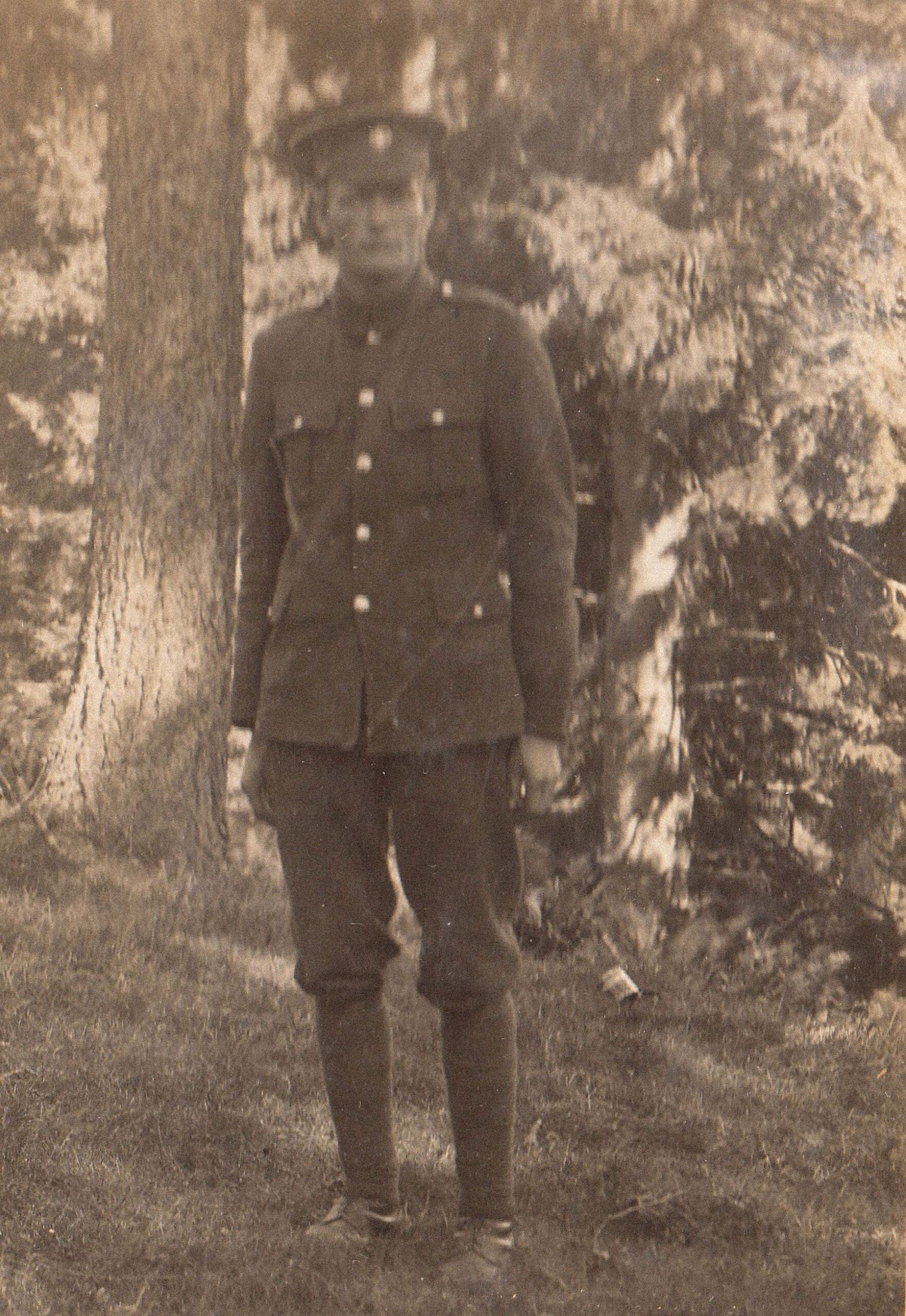 A sepia photograph of an unsmiling man in uniform and cap, standing in shadow on grass. Behind him is a stand of conifers in dappled light.