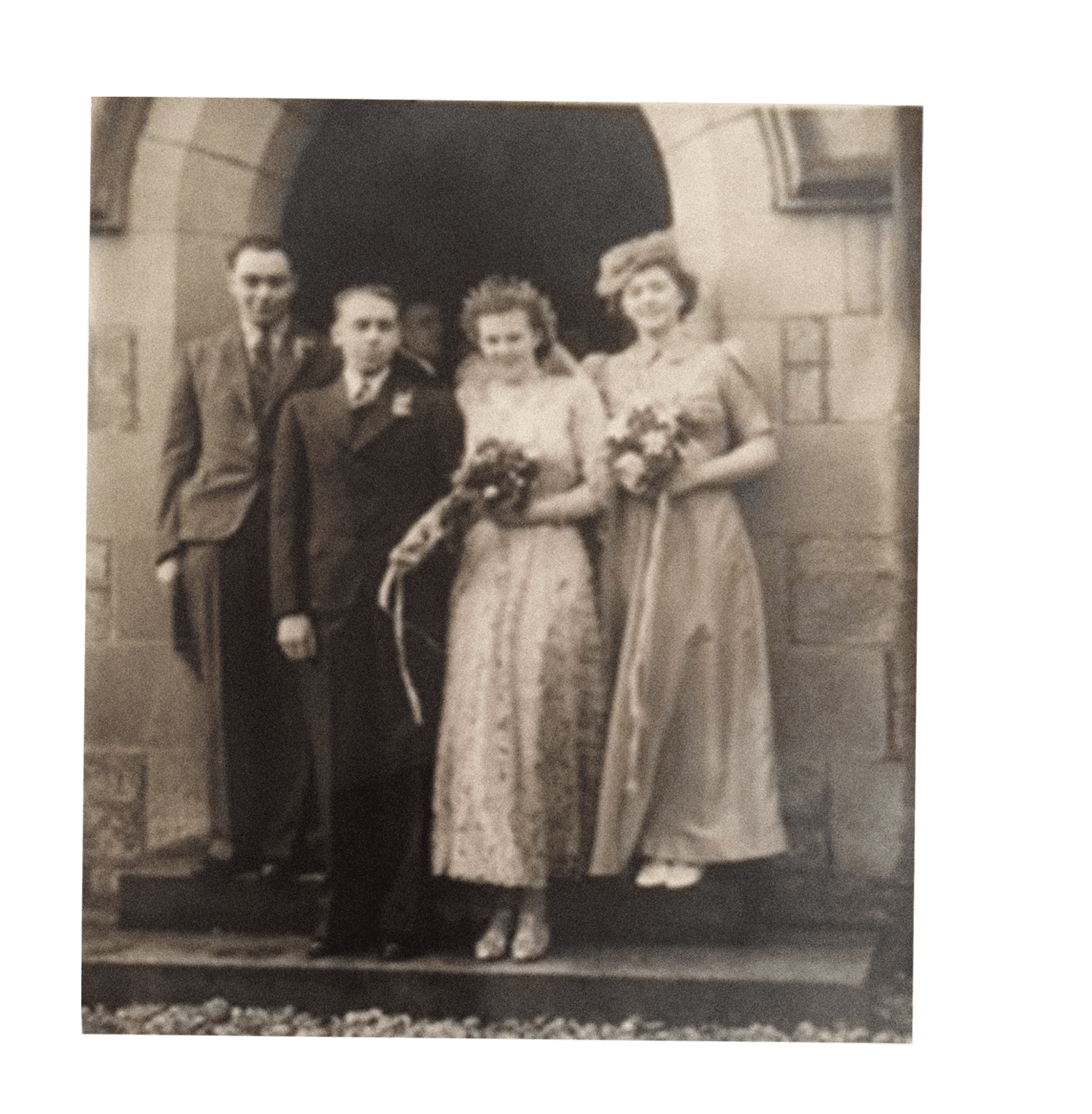 A sepia photograph of two men and two women standing just outside the arched portico of a stone building. The men are on the left, wearing suits and ties. The women, on the right, are wearing long gowns and holding bouquets.
