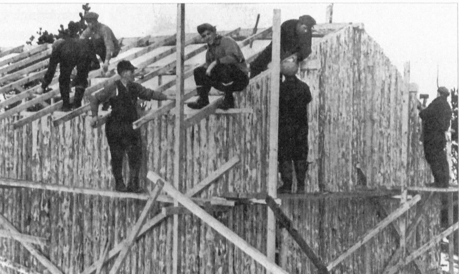 A black and white photo of a handful of men at work. They stand on a rough wooden scaffold or perch on the framed-in roof of a log cabin.