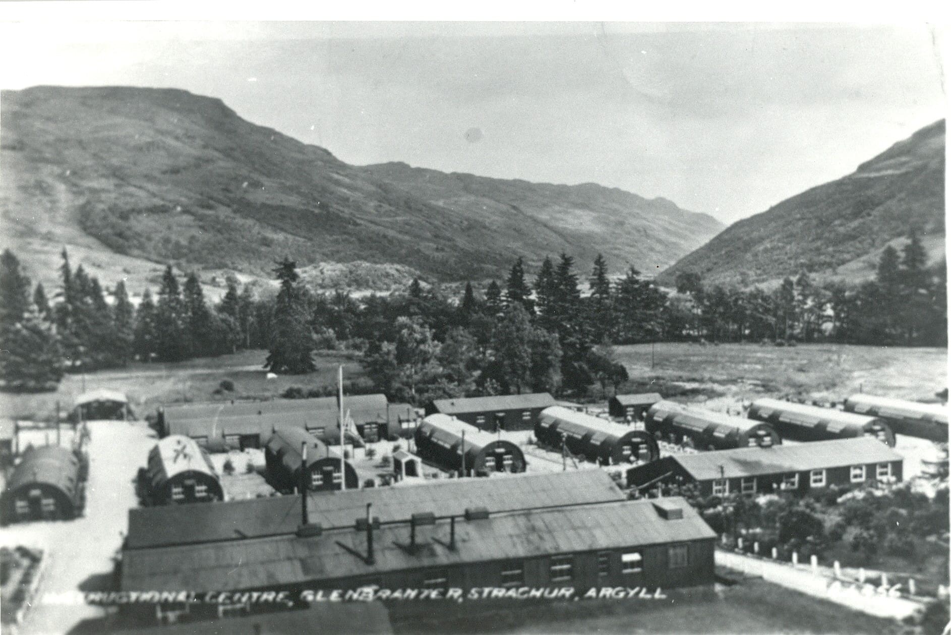 A black and white photograph of long low buildings grouped in neat rows on flat land. Behind them is a stand of trees. Valley sides recede in the distance.