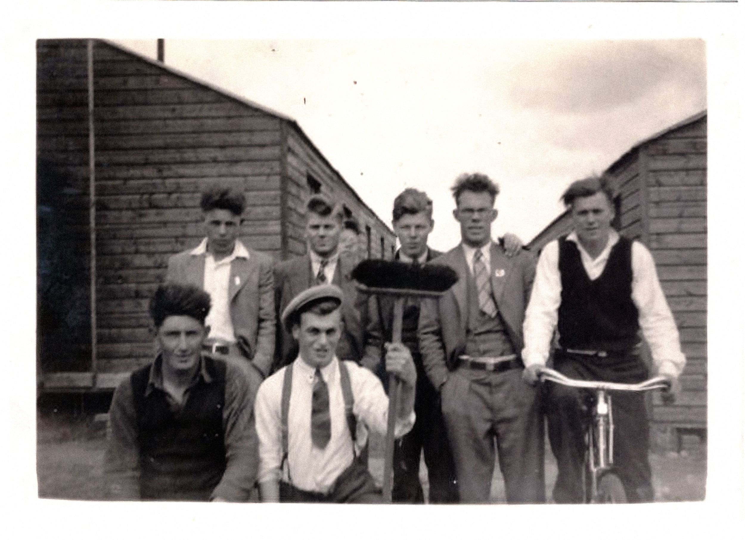 A black and white photograph of a handful of young men, some in shirts and ties, posing in the gap between a row of bunkhouses. One man sits on a bike, another holds a push broom.