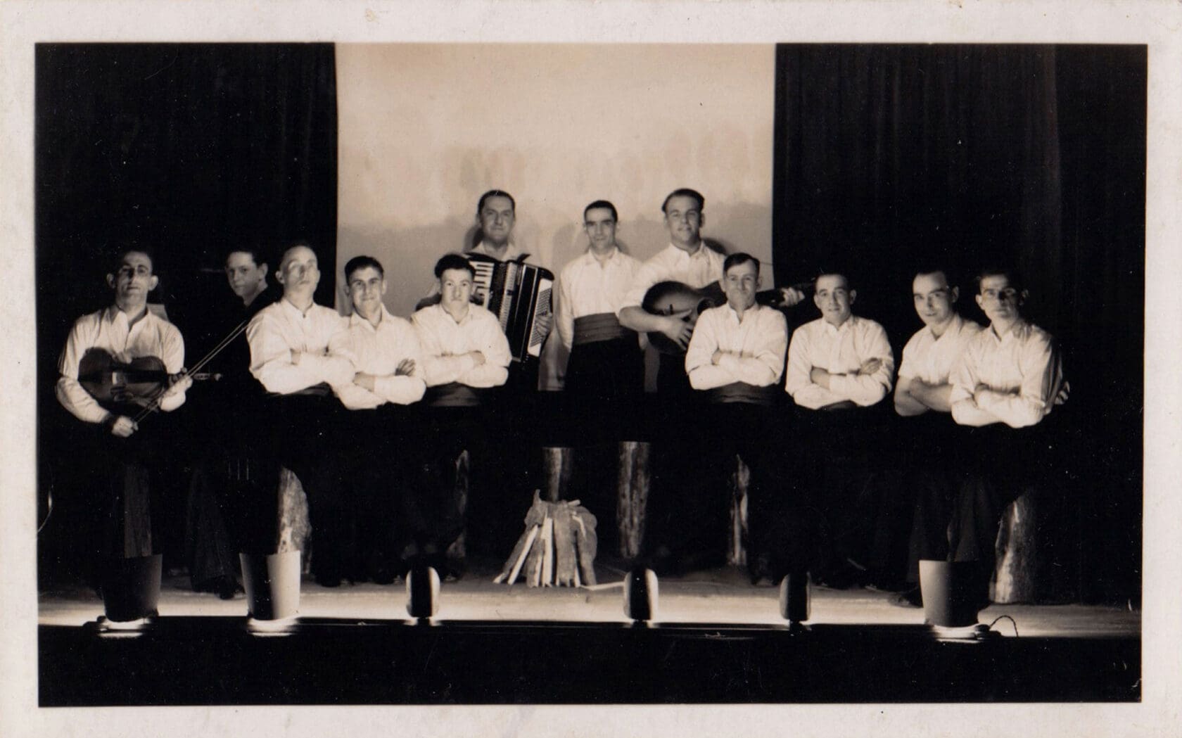 A black and white photograph of a row of eight men in white shirts lit by footlights that border a stage. A few more men stand behind them with a guitar and accordion.