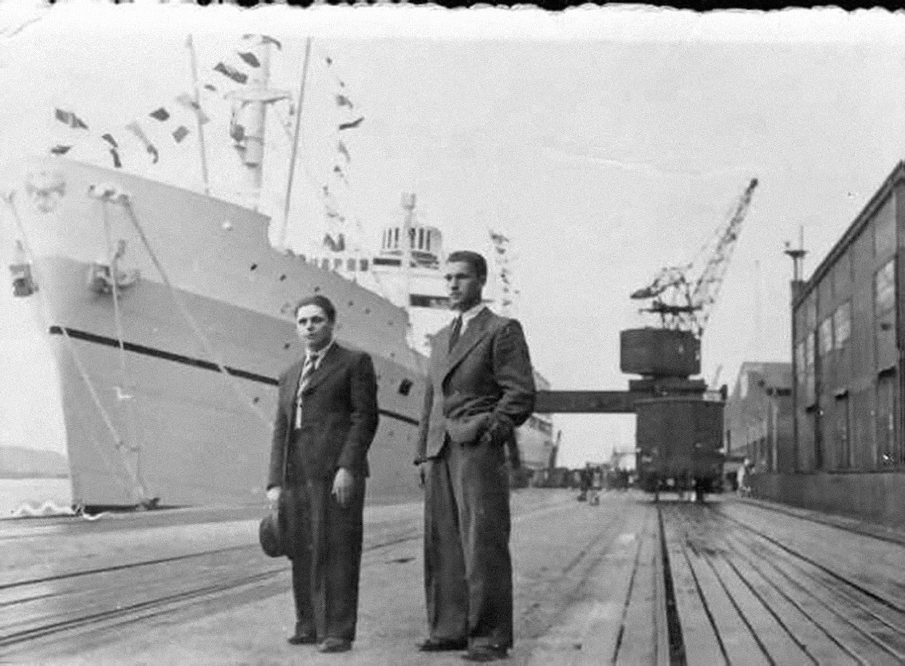 A black and white photograph of two men in suits and ties, standing near the bow of a white ship. A larger crowd and dock trams, buildings, and equipment can be seen in the background. Flags on fore and aft wires flutter on the ship.