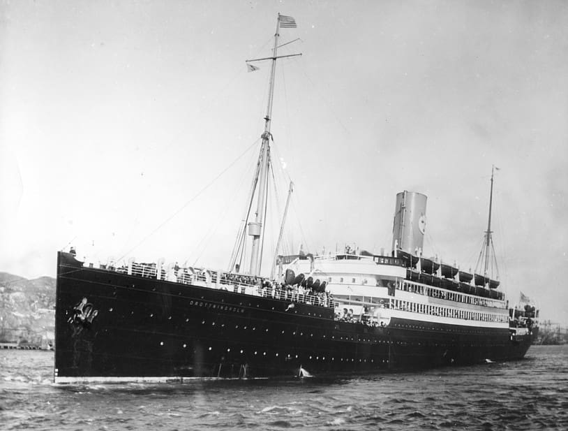 A black and white photo of the left (port) side of a ship with a dark hull and white superstructure, which is steaming toward the camera. Many people are visible on the forward deck and along railings outside the cabins. Two rows of portholes are visible along the ship’s hull. Land can be seen behind the ship.