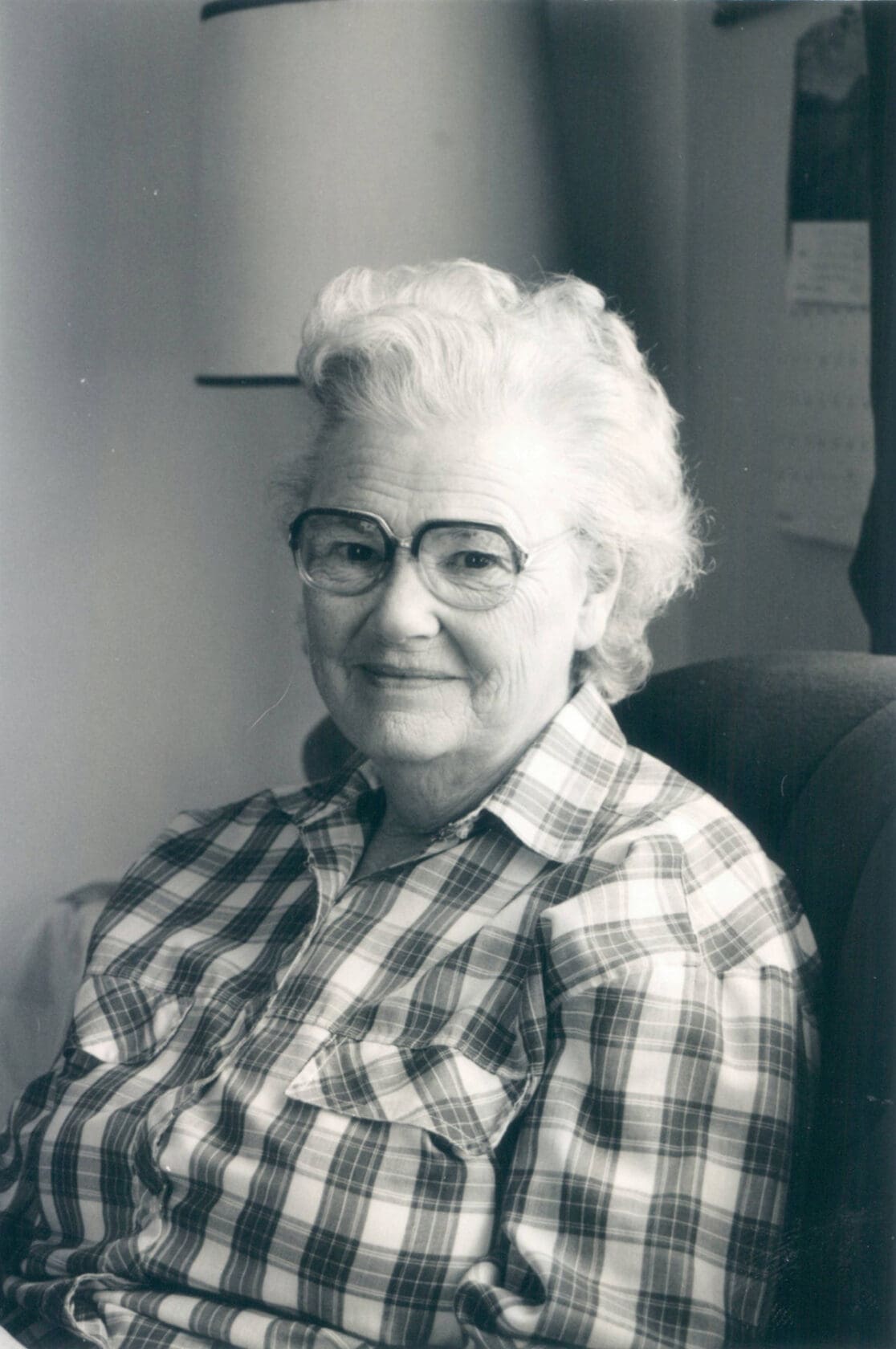 A black-and-white photo of a smiling white-haired woman wearing large glasses and a plaid shirt sitting in a dark chair. Her body is turned slightly and she looks directly into the camera, smiling.