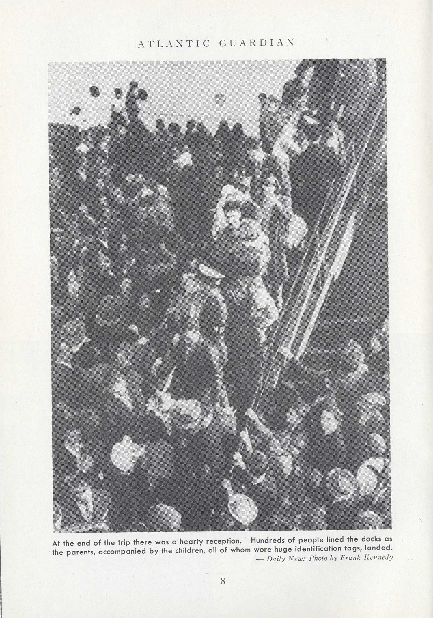 A black and white photograph, taken from above, shows a crowded gangplank that is surrounded by a throng of greeters. The caption reads: At the end of the trip there was a hearty reception. Hundreds of people lined the docks as the parents, accompanied by the children, all of whom wore huge identification tags, landed.
