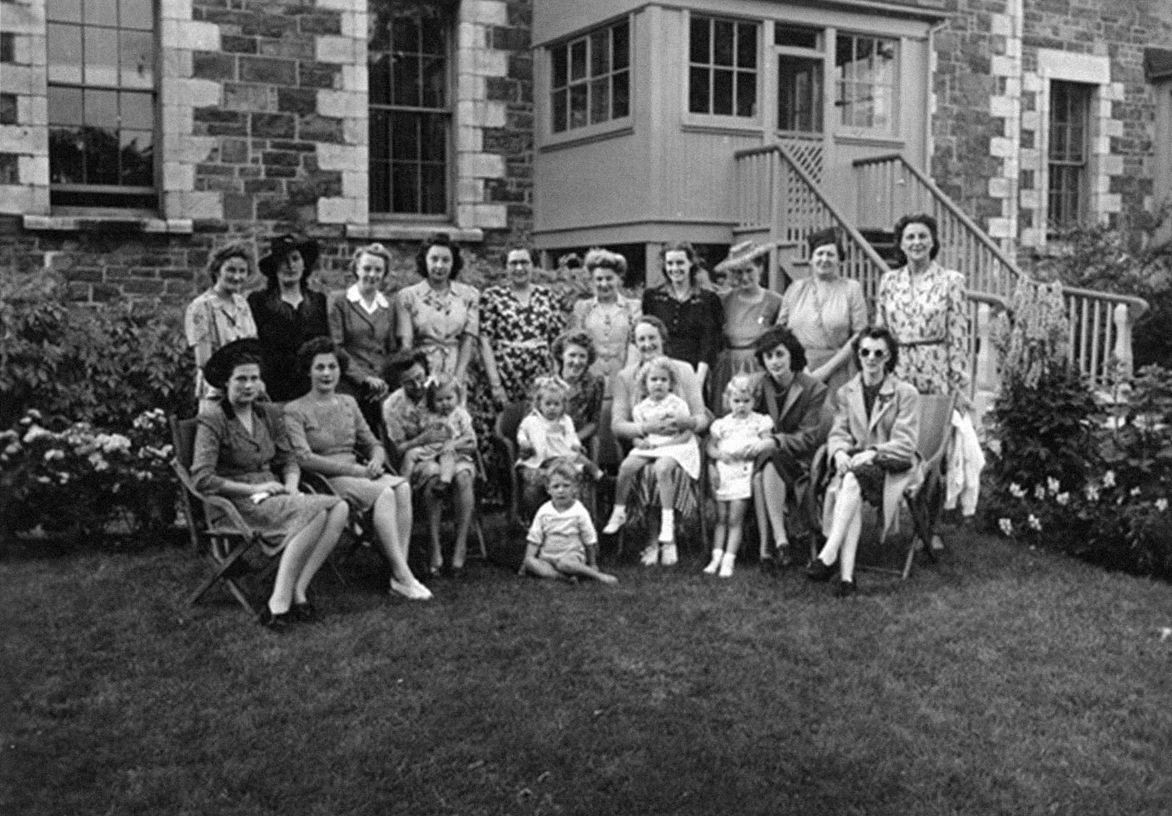 Seventeen women and a handful of children pose in the garden outside a stone building. Seven women are seated in a semi-circle, the others stand in a row behind them. All are wearing dresses and a few have hats and coats.